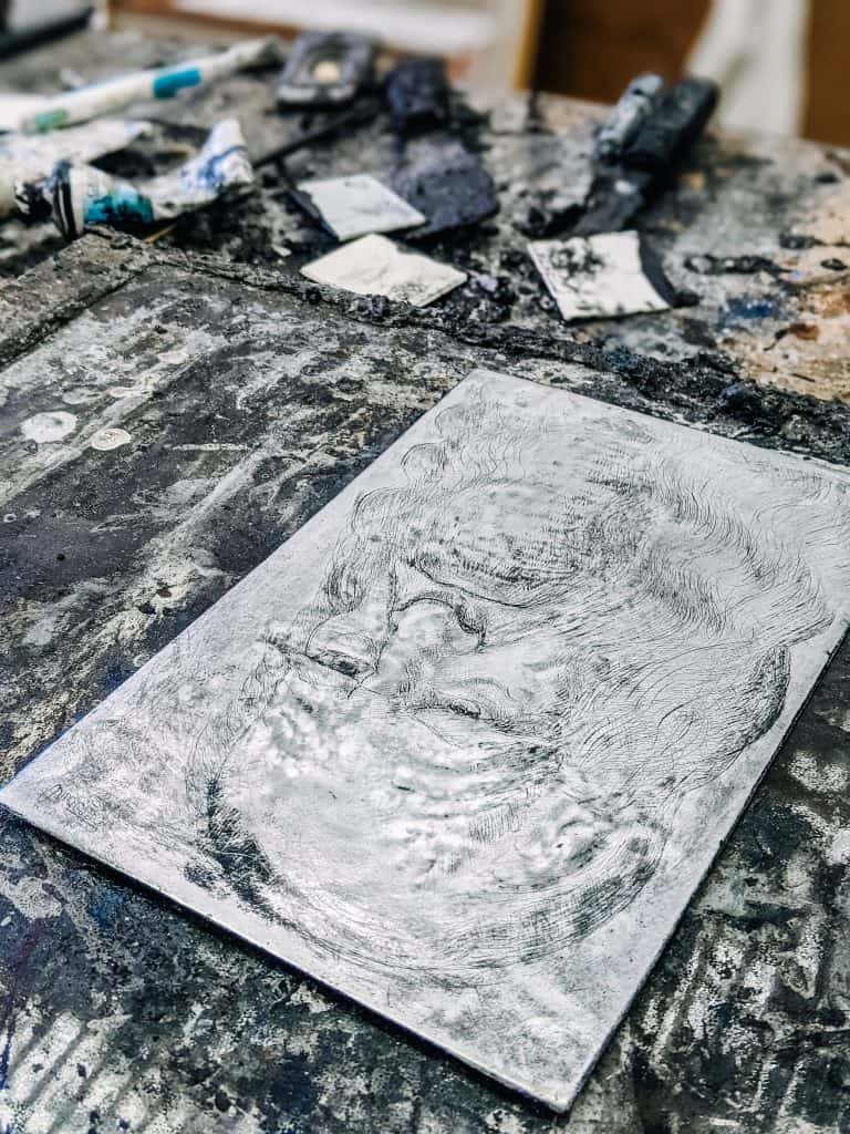 A textured, monochromatic artwork lies on a paint-splattered table in an artist's studio in Jerome Ghost Town. Surrounding the piece are various painting tools and supplies, adding to the creative and rustic ambiance of the space.