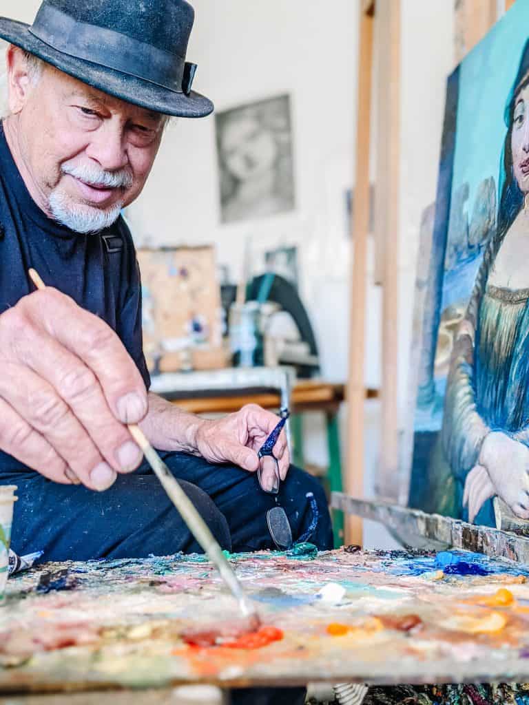 From the creative ambiance of Jerome Ghost Town. An elderly artist named Robin John Anderson with a white beard, wearing a black shirt and hat, paints in a studio filled with art supplies and canvases. One prominent canvas features a woman resembling the Mona Lisa.