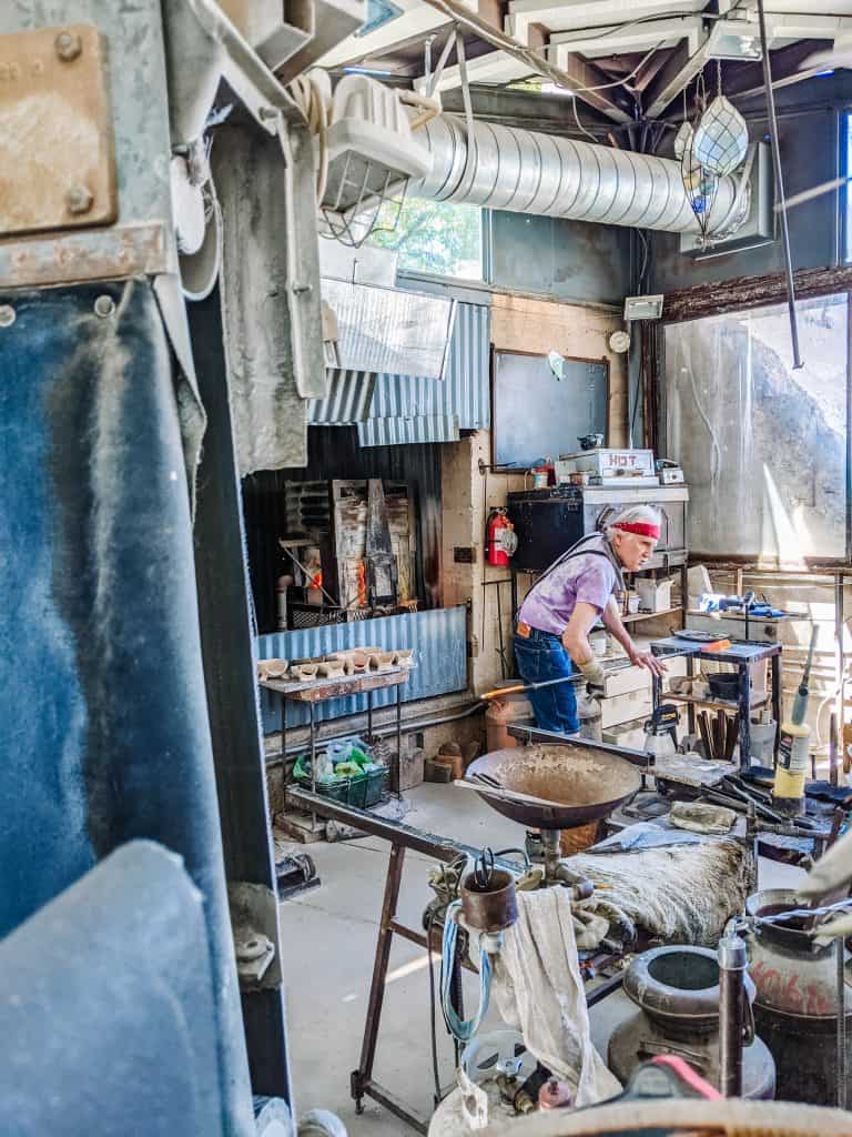 In Jerome Ghost Town, an artist in a red bandana and apron works intently in a cluttered, rustic studio filled with metalworking tools and equipment and a space, bathed in natural light.