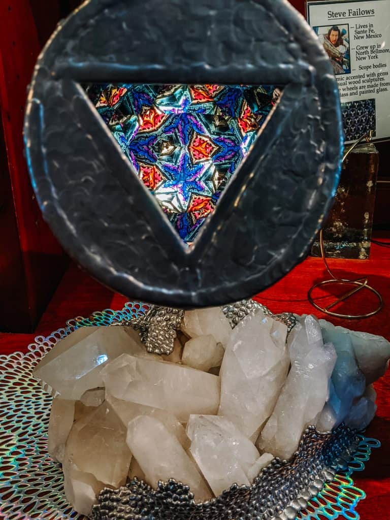 A vibrant kaleidoscope pattern is viewed through a circular metal frame, set above a cluster of white quartz crystals on a mesh surface. The background includes an informational sign about the artist, adding a touch of local history of the Jerome Ghost Town.