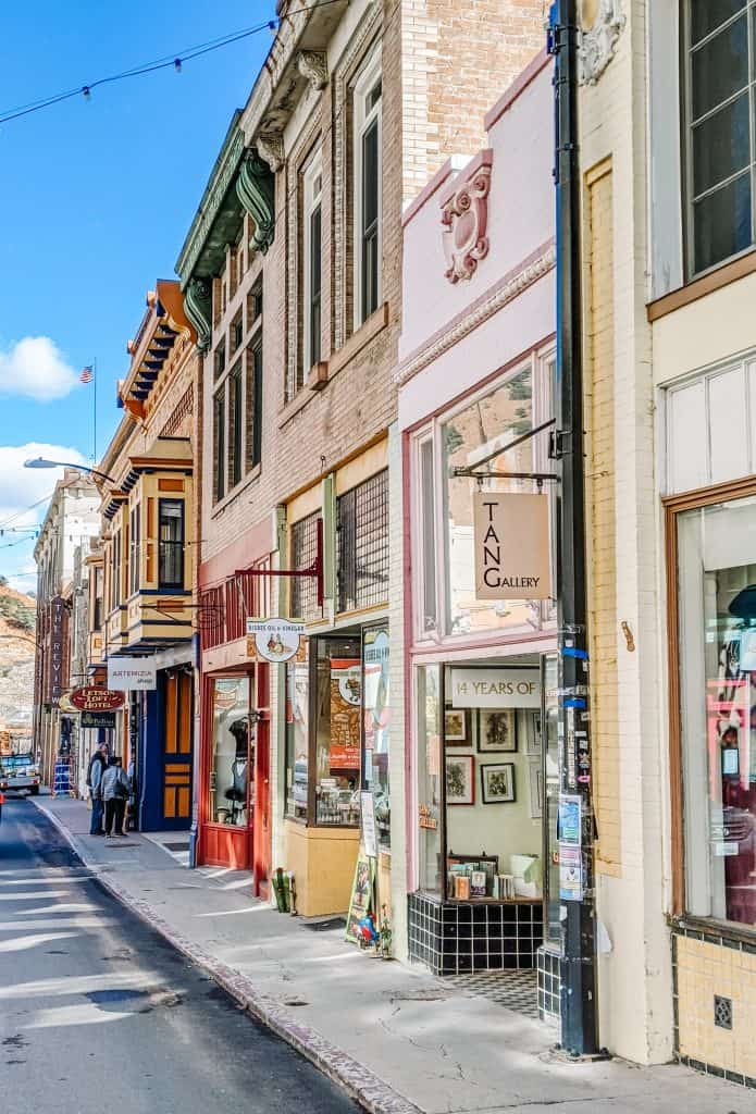 Gorgeously painted colors from the shops in Bisbee City Arizona, with this street's colorful storefronts and historic architecture. The T.A.N. Gallery stands out with its artistic display, celebrating "14 years of Art." The vibrant atmosphere of downtown Bisbee, set against a clear blue sky.