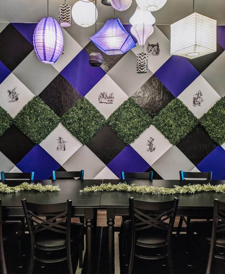 A dining area featuring a black and white checkered wall with purple accents and greenery. The long black table is surrounded by chairs and decorated with garlands. Overhead, colorful lanterns hang, adding a magical touch to the atmosphere of Drink Me Tea Room.