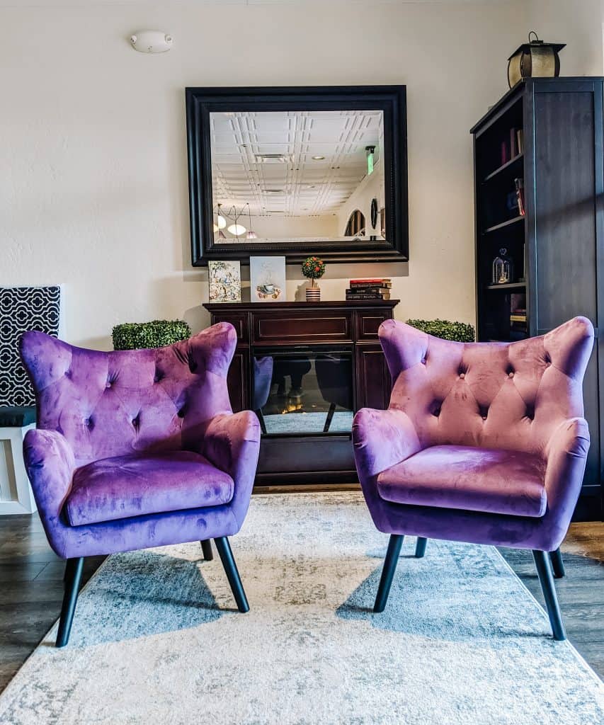 This cozy seating area featuring two plush purple chairs with tufted backs at Drink Me Tea Room, arranged on a soft rug in front of a large mirror, creating an inviting spot to relax and enjoy tea.