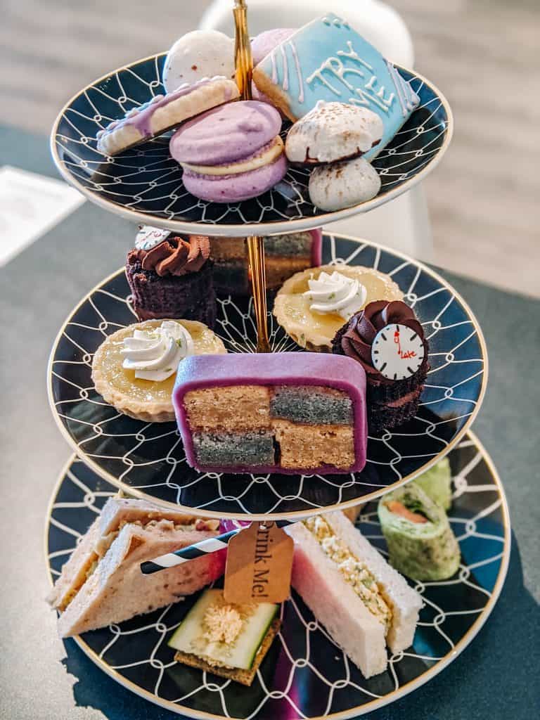 A tiered tray filled with a variety of treats. The top tier holds colorful macarons and delicate meringues. The middle tier features mini tarts, cupcakes, and a slice of lively layer cake. The bottom tier is arranged with an assortment of finger sandwiches, each with unique fillings, perfect for a classic tea time at Drink Me Tea Room in Arizona.