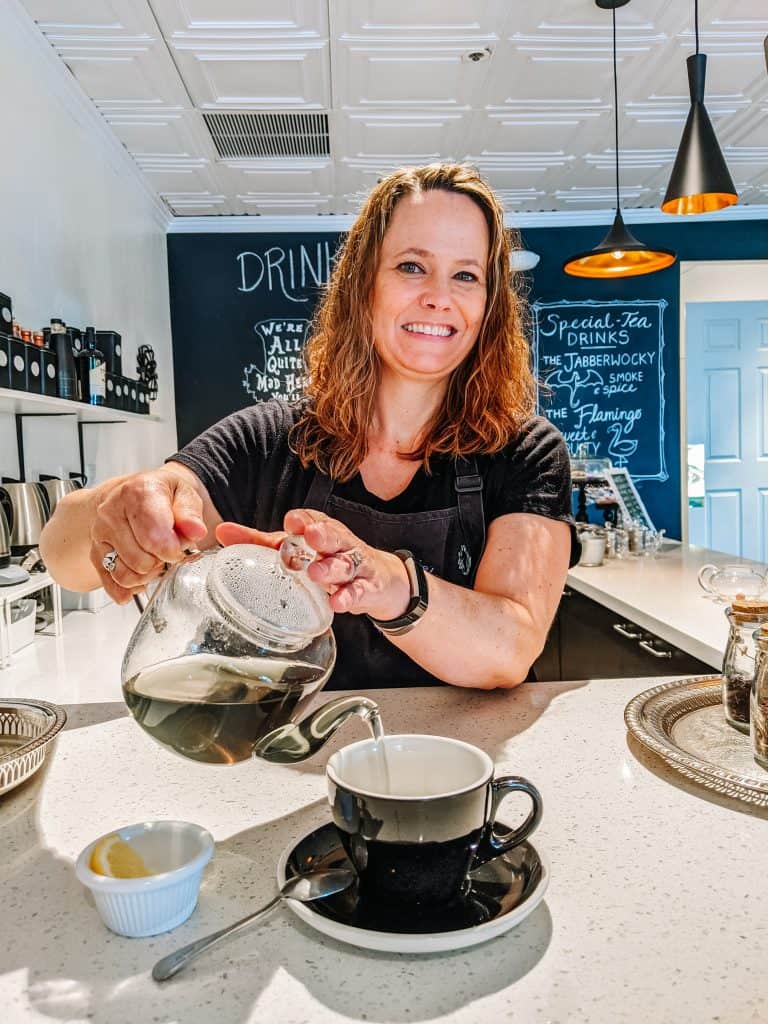 Melissa Harlan the owner of Drink Me Tea Room, pouring tea from a glass teapot into a black cup. She’s smiling warmly, creating a welcoming atmosphere with a background features the menu on a chalkboard wall,