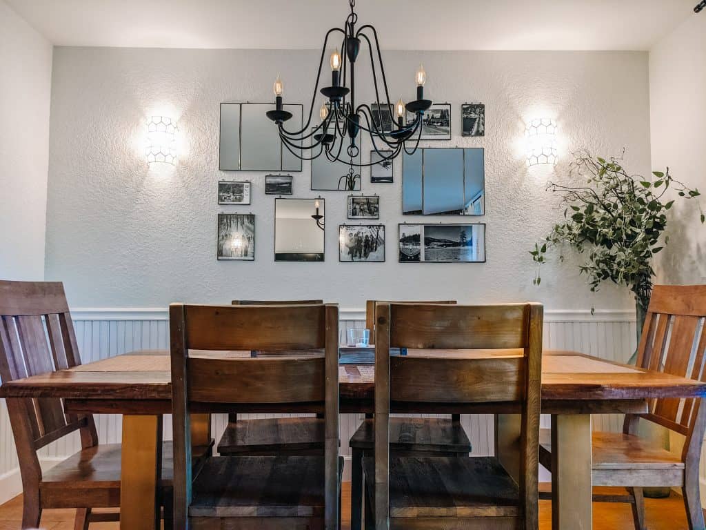 This image features a cozy dining room in the City of Big Bear, with a rustic wooden table and chairs set under a classic black chandelier. The wall is decorated with a collection of mirrors and black-and-white photos, adding a touch of charm and character to the space. A lush green plant in the corner brings a bit of nature indoors.