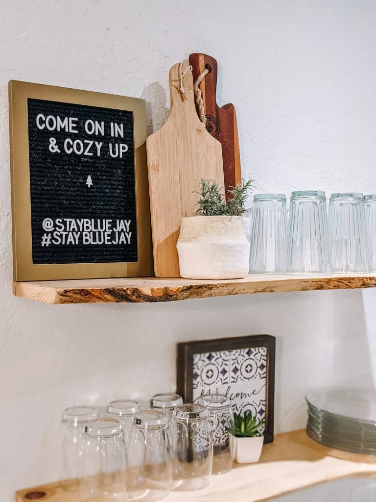 A wooden shelf adorned with a charming sign that reads, "Come on in & cozy up" in the City of Big Bear. Next to it, there's a cutting board, a small plant, and a row of neatly arranged glasses and plates. This welcoming and homey vibe is just a taste of the inviting atmosphere you'll find throughout the City of Big Bear.