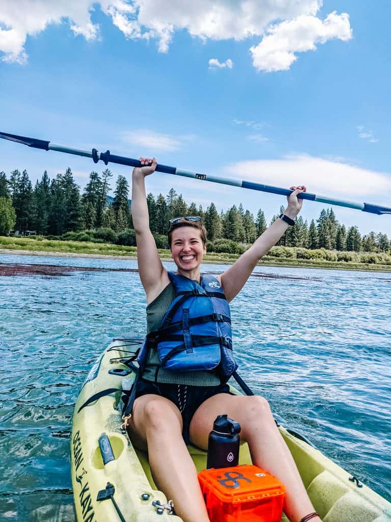 Kayaking on Big Bear Lake, raising my paddle wearing a blue life vest and a big smile on my face. The City of Big Bear offers an amazing experience on the water with stunning views of lush trees and clear blue skies.