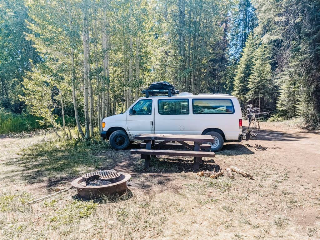 Here is my van parked in a forest campsite, surrounded by tall trees and natural beauty. The picnic table and fire pit are ready for a cozy evening under the stars. Van traveling in such peaceful spots.