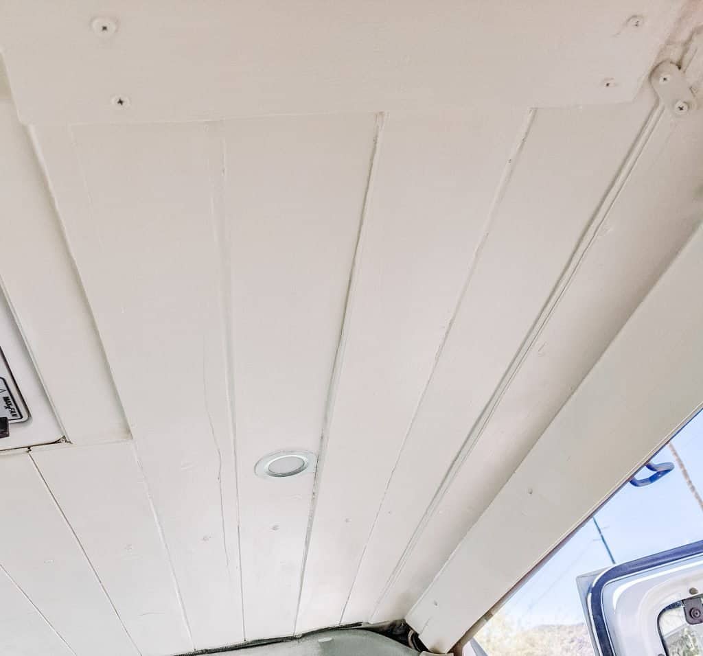 The Ford camper van ceiling is covered with neatly aligned white wooden planks, featuring a small recessed light in the center for illumination.