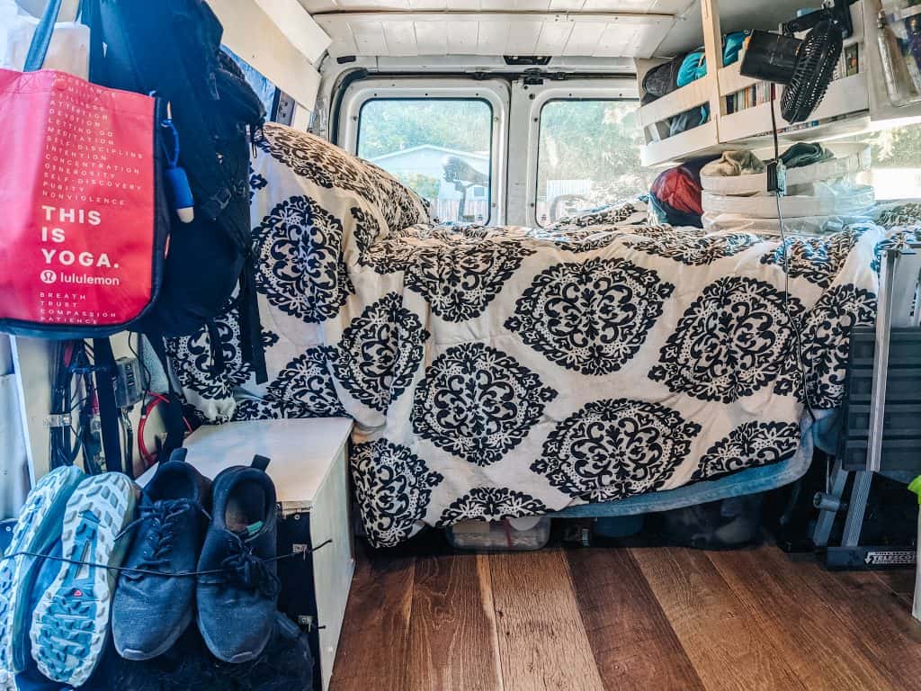 Inside a camper conversion van, this cozy setup features a bed with a black and white patterned blanket, neatly organized storage shelves, and essential travel gear like shoes and bags, all creating a functional and comfortable living space on the road.