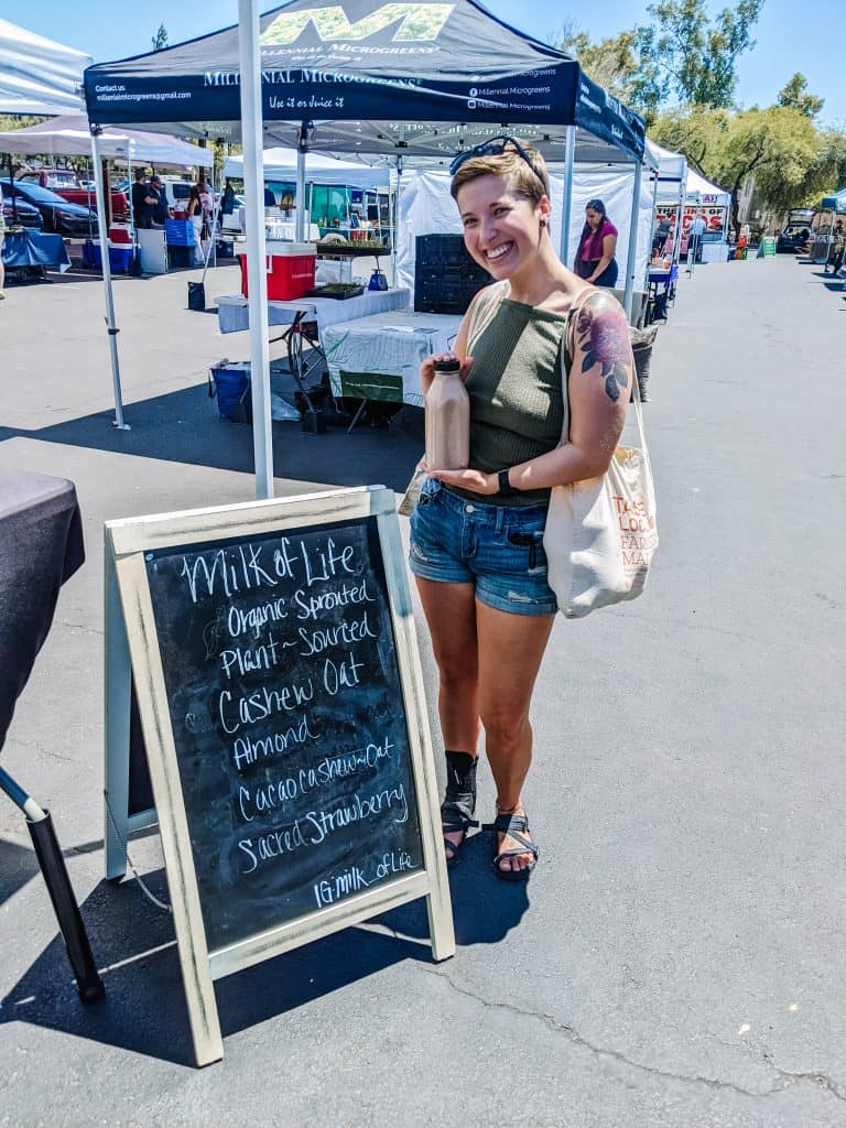 Standing at a farmers' market holding a glass bottle of milk, next to a chalkboard sign that lists various plant-based milk options. This highlights a practical way of how to help the environment