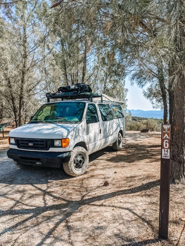 A white van is parked at a campsite surrounded by tall trees, with a signpost labeled "A6" indicating the site number, capturing the essence of traveling in a van amidst nature.