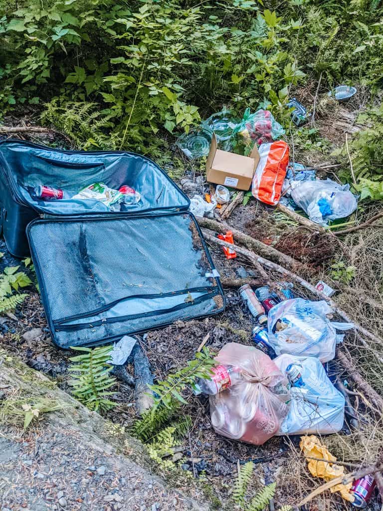 A pile of litter, including an open suitcase, plastic bags, bottles, and food wrappers, discarded in a forest area. Traveling in a van means you should always be mindful of leaving no trace and keeping natural spaces clean to protect the environment.