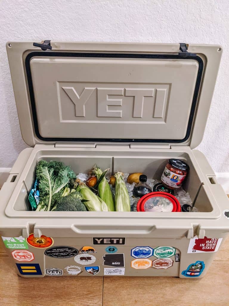 A YETI cooler filled with fresh vegetables and various food items is open and ready for a trip. The cooler is decorated with multiple colorful stickers and is essential for keeping food fresh in my converted van.