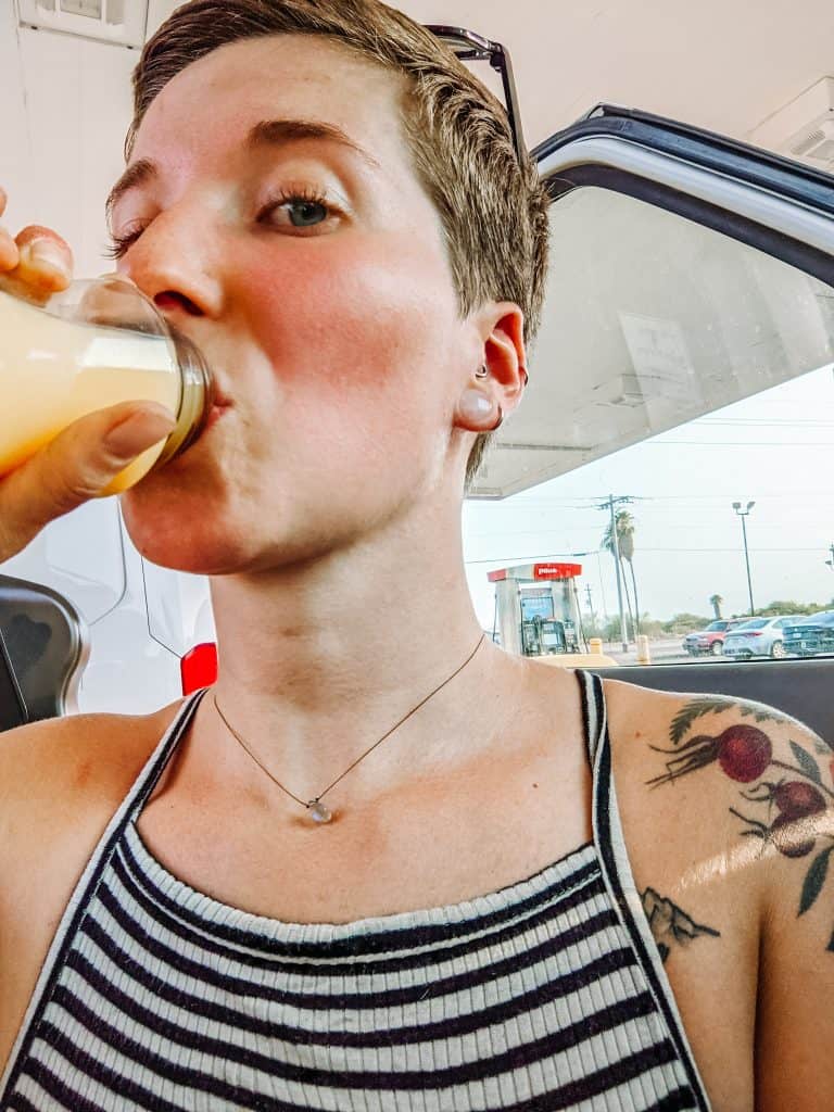 Drinking juice while standing next to my converted van. The open door of the van reveals a glimpse of the interior, and a gas station can be seen in the background.