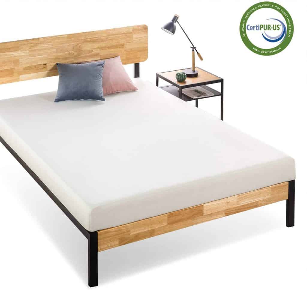 A neatly made bed with a wooden headboard, two pillows, and a CertiPUR-US label sits next to a small side table with a lamp and plant, perfect for a camper conversion van.