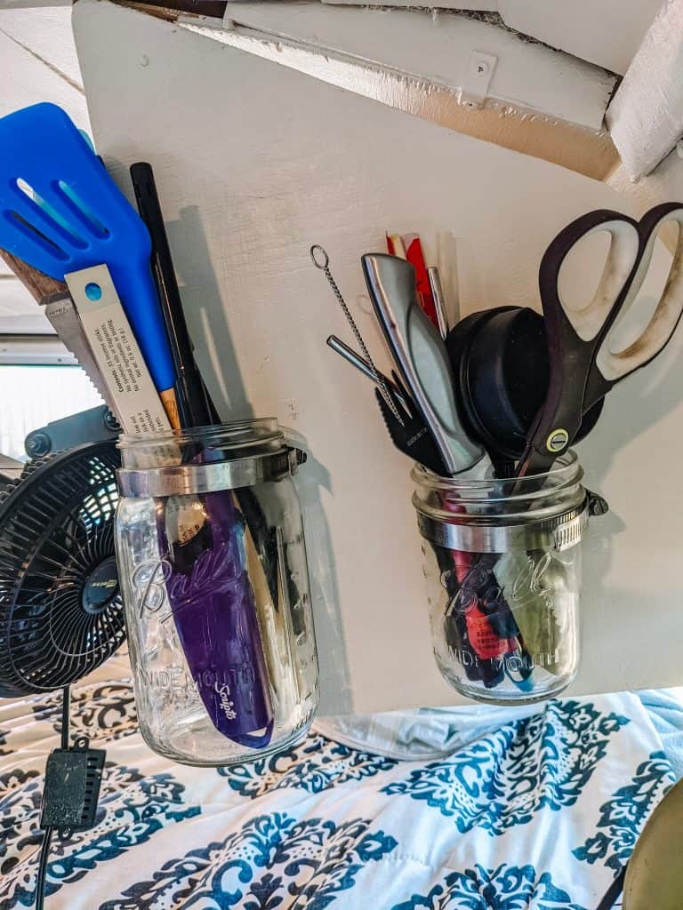 In this camper conversion van, two glass jars mounted on a white wall hold essential utensils like scissors, a spatula, and pens, making efficient use of vertical space for easy access and organization.