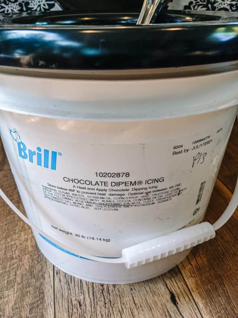 A large white container with a black lid labeled "Brill" containing chocolate dip icing, which has been repurposed as a portable toilet for campervan life. The container's sturdy build and handle make it a practical choice for van dwellers needing a compact and durable toilet solution.