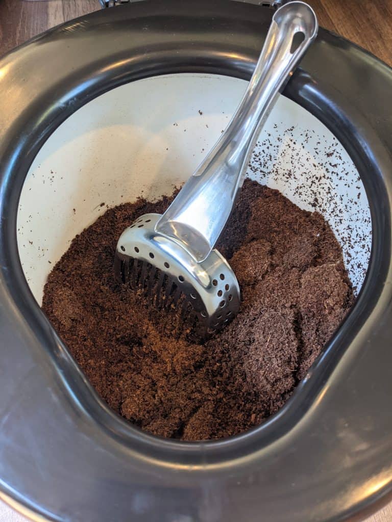 The interior of a portable toilet for campervan life, filled with a brown, soil-like substance. A metal scooper rests inside, ready for use. The setup demonstrates an eco-friendly, composting method for waste management.