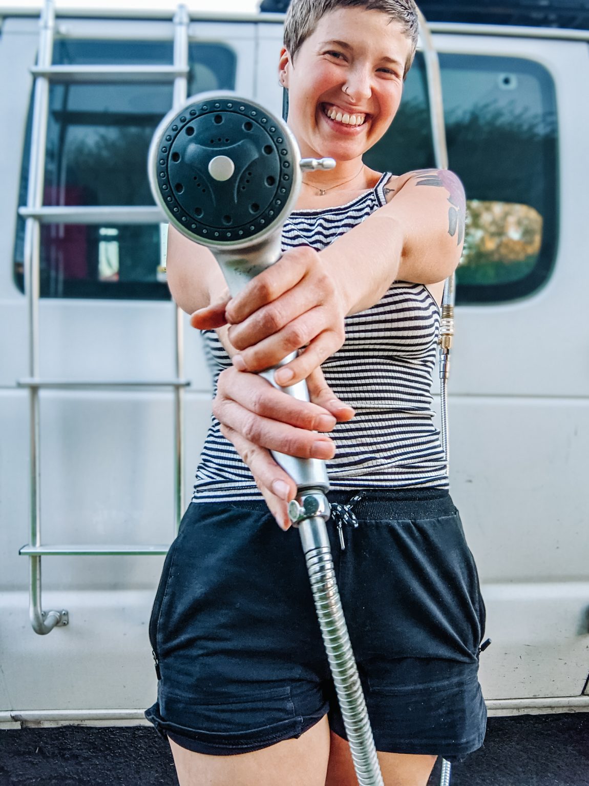 I am standing in front of my camper conversion van, smiling and holding out the showerhead, ready to use the convenient outdoor shower setup.