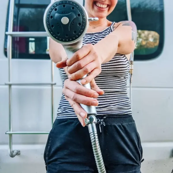 I am standing in front of my camper conversion van, smiling and holding out the showerhead, ready to use the convenient outdoor shower setup.