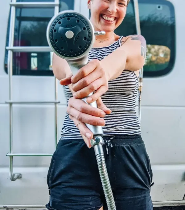 I am standing in front of my camper conversion van, smiling and holding out the showerhead, ready to use the convenient outdoor shower setup.