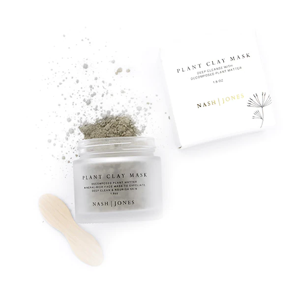 A jar of Nash and Jones Plant Clay Mask is open with some clay powder spilled out next to a small wooden spoon, alongside the product's white box with minimalist design, making it a great option for zero waste gifts.