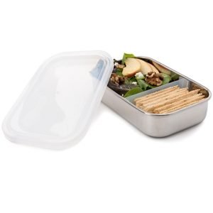 A stainless steel container with a clear plastic lid, partially open to reveal a selection of snacks you can bring on an airplane, including sliced apples, leafy greens, and crackers.