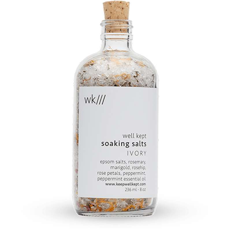 A clear glass bottle with a cork stopper contains well kept soaking salts, making it an ideal choice for zero waste gifts. The label lists ingredients like epsom salts, rosemary, marigold, rosehip, rose petals, and peppermint essential oil.