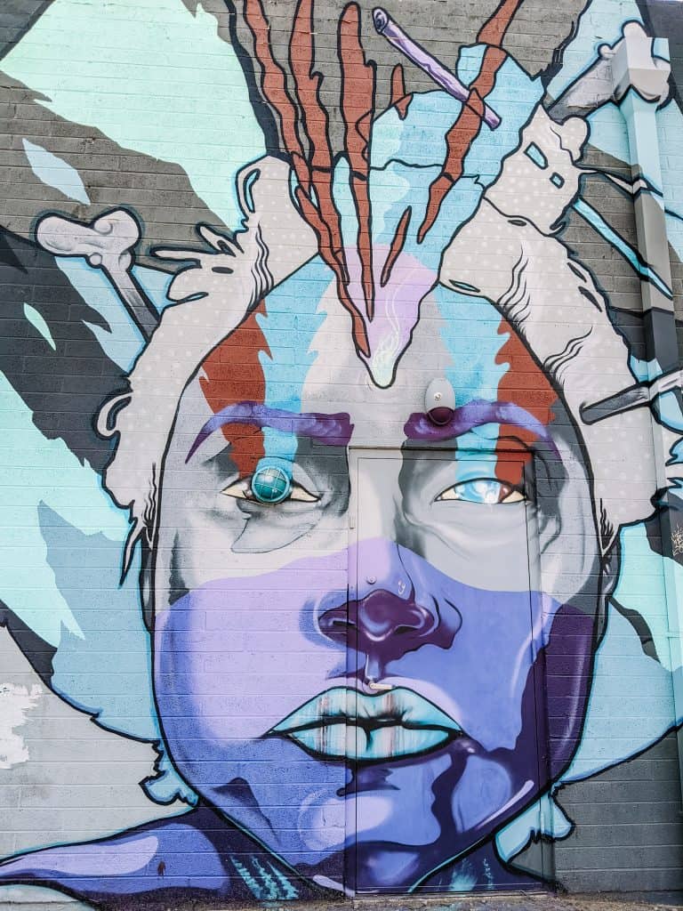 This murals in Phoenix features a bold and colorful portrait of a woman's face, with striking blue, purple, and red hues, and abstract elements surrounding her head, adding depth and intrigue.