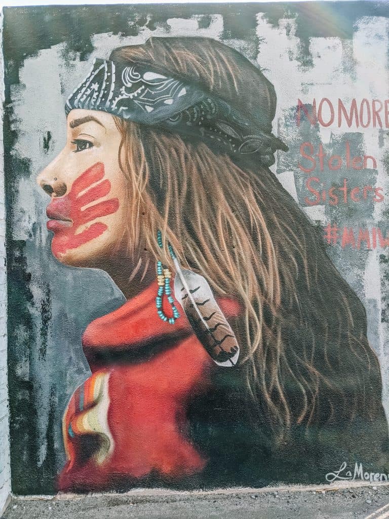 This mural in Phoenix shows a Native American woman with a red handprint over her mouth, symbolizing the issue of missing and murdered Indigenous women. She is wearing a headscarf and feather earrings, with the words "No More Stolen Sisters" in the background, highlighting the powerful message and cultural significance of this piece.