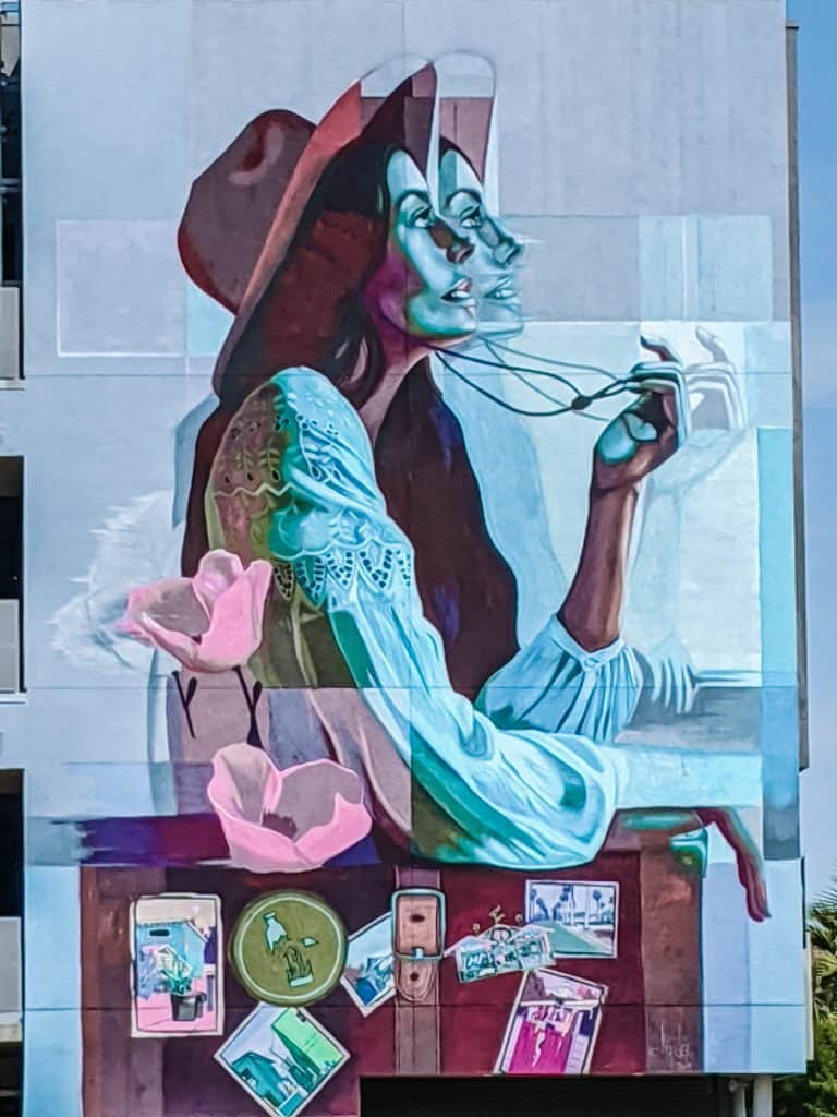 Features a large murals in Phoenix, showing a woman in a hat with overlapping images of her face, giving a sense of motion. She is holding her glasses, with vibrant colors and details like flowers and various objects on a table below her. The mural adds a dynamic and artistic touch to the building's front.