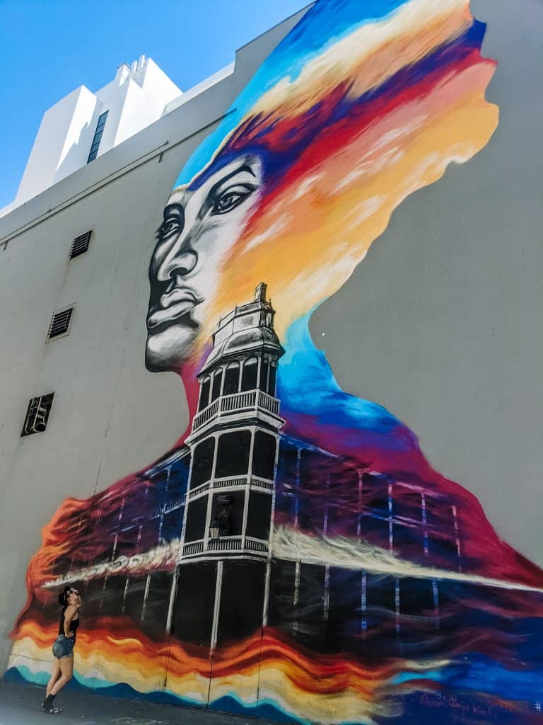 I'm standing on the side of a vibrant murals in Phoenix, shows a woman's face emerging from a colorful explosion of reds, blues, and oranges, with a black and white tower structure below her.