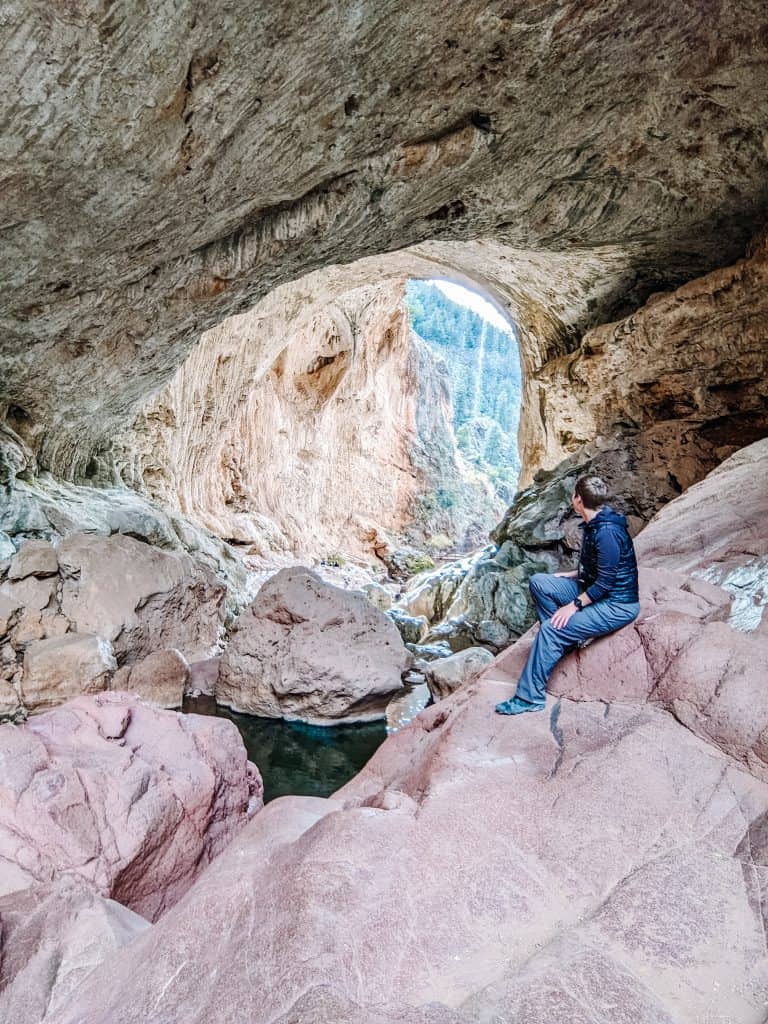 I'm sitting on a large rock inside a massive cave with a stunning view of Tonto Natural Bridge, surrounded by rocky formations and a small pool of water below.