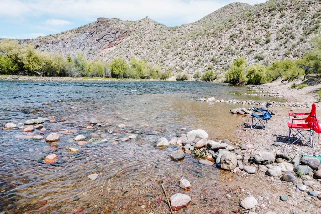 A peaceful riverside spot after an Arizona hiking trip, with two empty camping chairs set up on the rocky shore, surrounded by clear water, green trees, and hills under a bright blue sky.