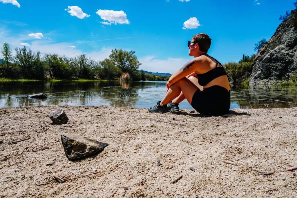 Sitting by a tranquil river after an Arizona hiking trip, I'm wearing a black sports bra and shorts, enjoying the sunny day with green trees and a clear blue sky in the background.