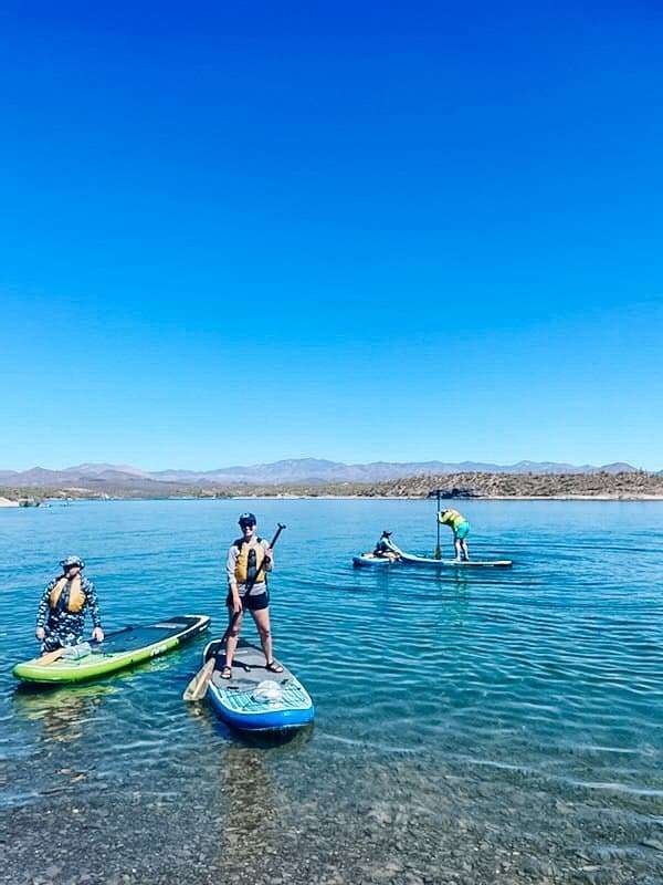 In this image from our adventure guide, I'm in the middle paddleboarding on a clear blue lake with three people in the background, all wearing life vests and enjoying the sunny day with mountains visible in the distance.