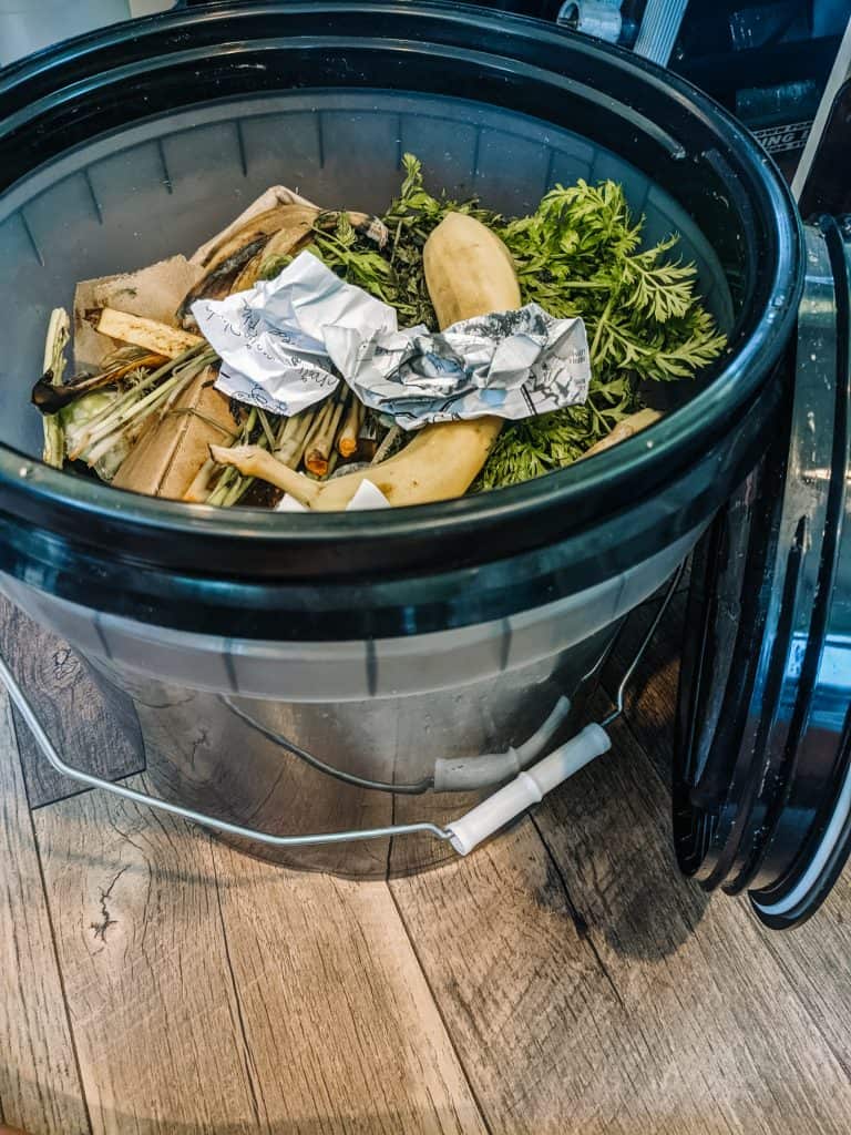 A compost bin filled with kitchen scraps, including vegetable peels, carrot tops, and a piece of aluminum foil, illustrating what is compost.