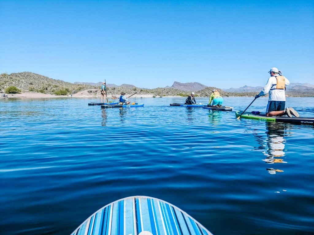 A group of paddleboarders on a calm blue lake with a striped paddleboard in the foreground, surrounded by hills and mountains under a clear blue sky, perfect for an adventure guide.