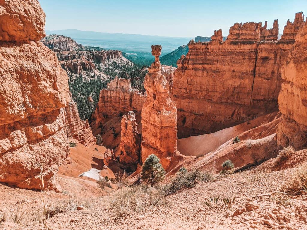 The stunning rock formations of Bryce National Park, featuring tall, narrow spires called hoodoos that rise dramatically from the canyon floor. The red and orange hues of the rock contrast beautifully with the distant green forest and blue sky