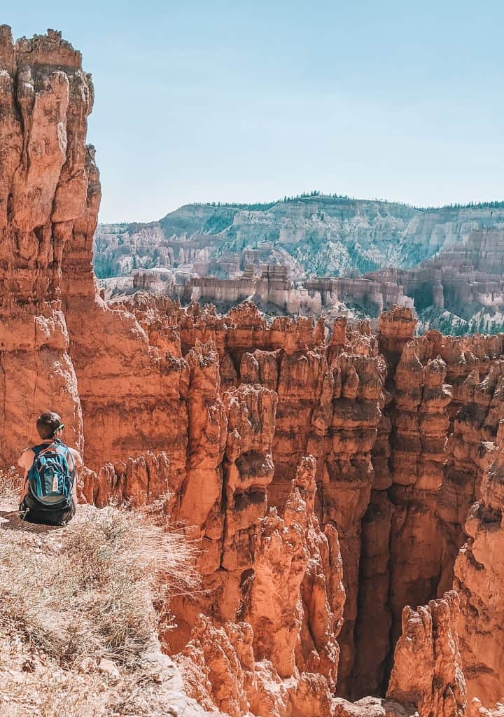 That's me sitting at the edge of a cliff in Bryce National Park, wearing a backpack and looking out over the stunning orange and red hoodoos and cliffs under a clear blue sky.