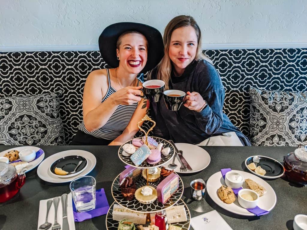 At Drink Me Tea Room me and my friend seated at a table filled with delicious pastries and treats, holding our teacups and smiling. The patterned black and white background and cozy cushions add to the inviting atmosphere.