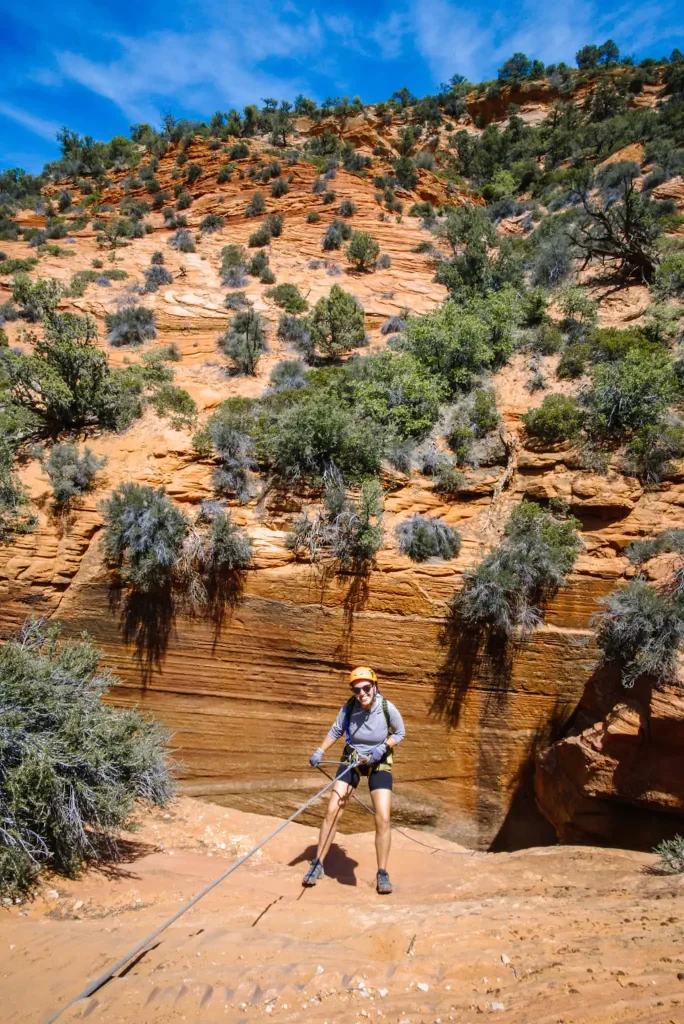 I'm rappelling down a rocky cliff at Zion Ponderosa Ranch Resort, surrounded by vibrant red and orange canyon walls and green shrubs, enjoying an adventurous day under a bright blue sky.