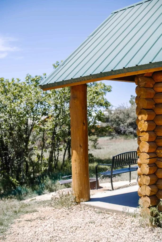 A cozy cabin with a green metal roof and wooden log walls at Zion Ponderosa Ranch Resort, surrounded by lush trees and greenery, with a black metal bench on the porch.