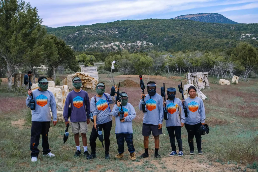 A group of people wearing matching gray shirts and protective masks are ready for a game of paintball at Zion Ponderosa Ranch Resort, set against a backdrop of lush green hills and a clear sky.