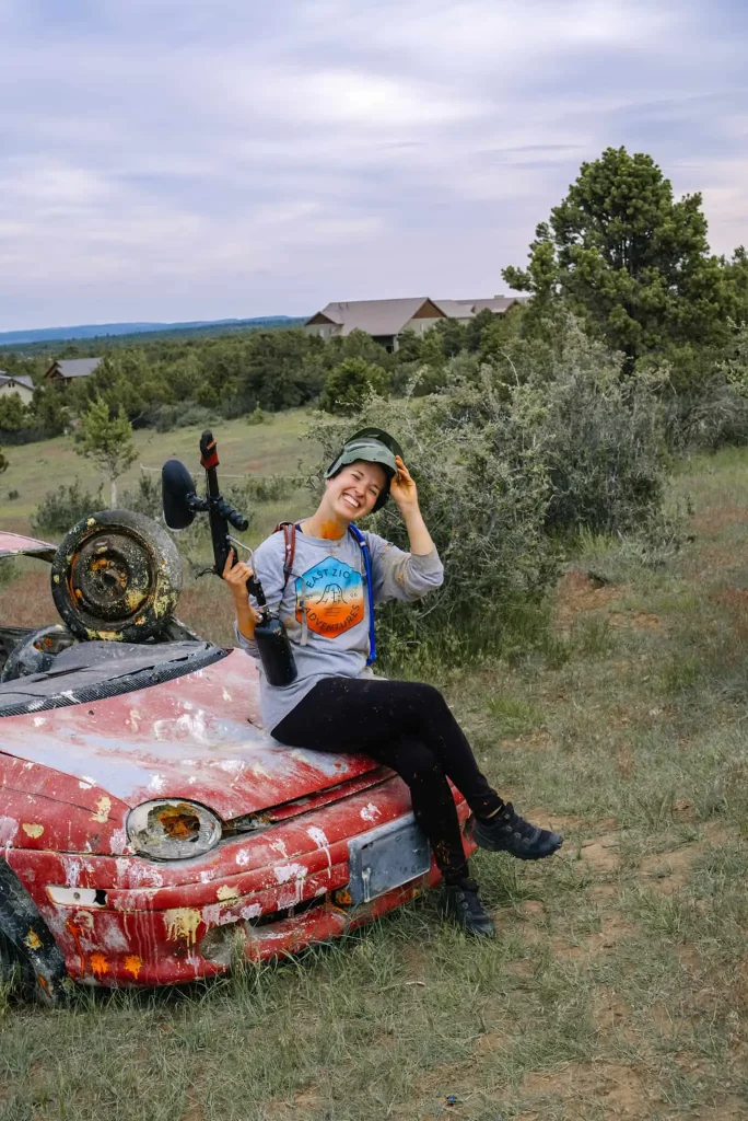 I am sitting on a paint-splattered red car, holding a paintball gun and smiling after a thrilling paintball game at Zion Ponderosa Ranch Resort. The background features the resort's cabins nestled among trees under a cloudy sky.