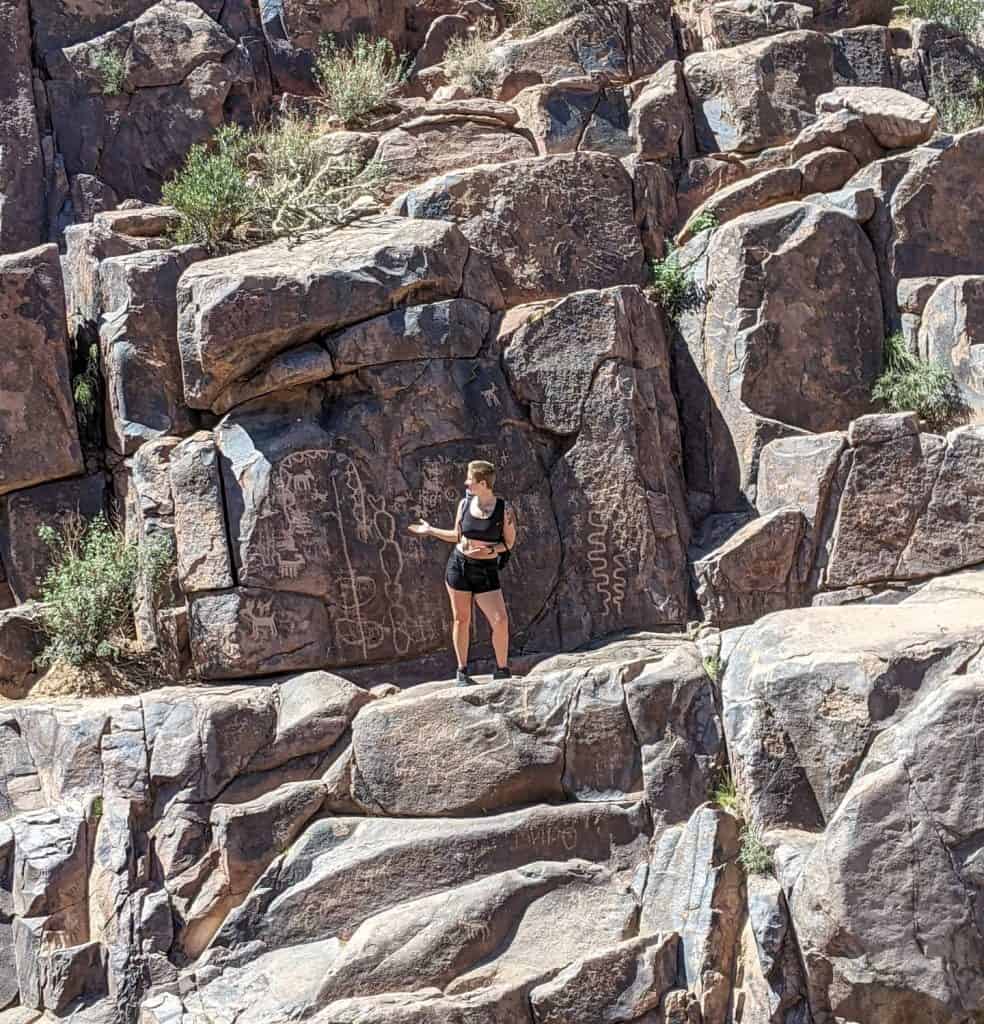 While hiking in Phoenix, I stood on a rocky ledge surrounded by large boulders etched with ancient petroglyphs, wearing a black tank top and shorts, taking in the rich history and stunning desert landscape.