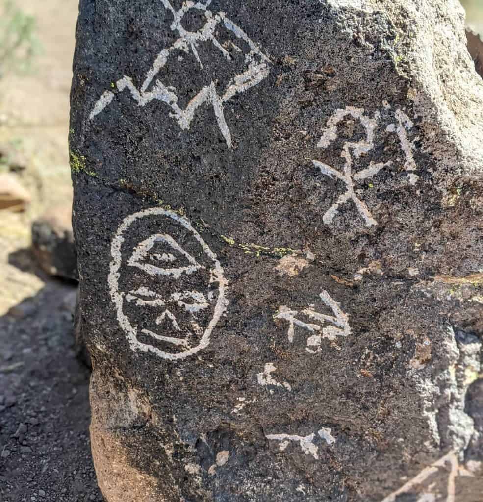 This image captures ancient rock carvings etched into a dark rock, featuring a human-like face and various symbols, which can be seen while hiking in Phoenix.