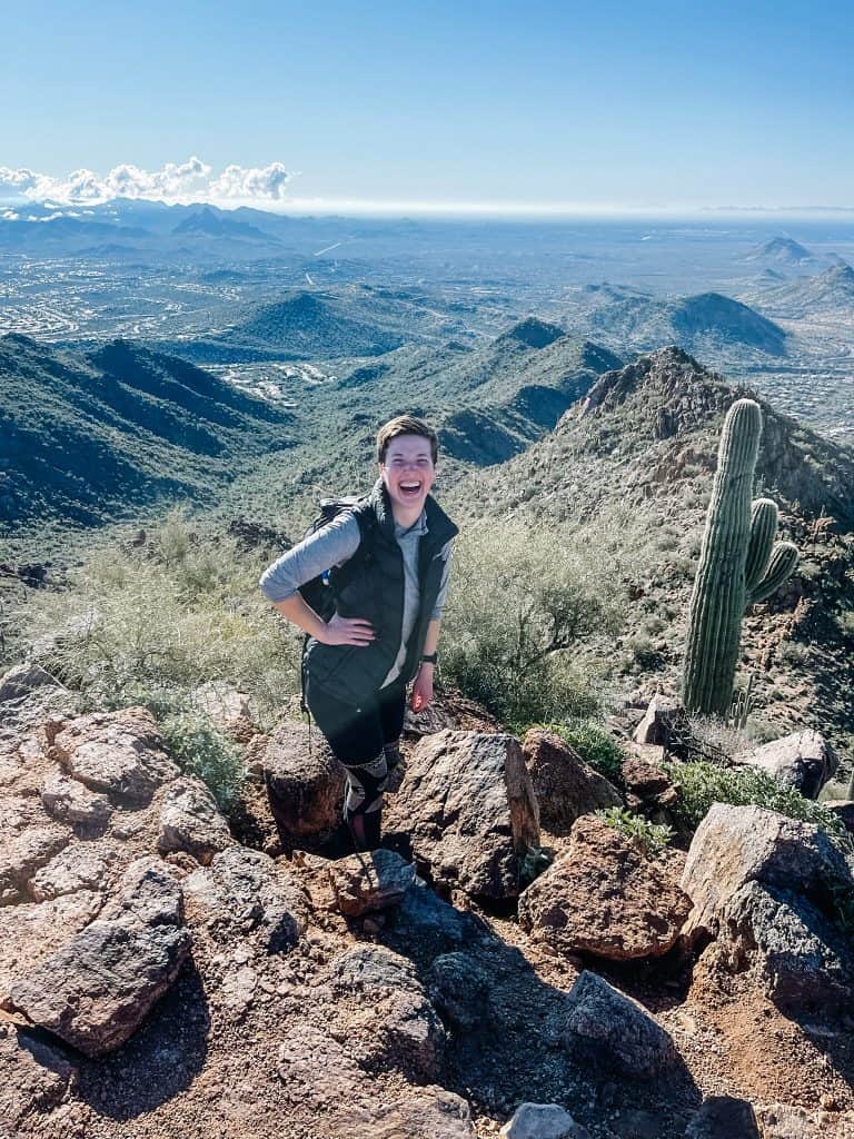 Here I am hiking in Phoenix, standing on a rocky outcrop with a vast desert landscape and distant mountains stretching out behind me, under a clear blue sky with a tall cactus nearby.